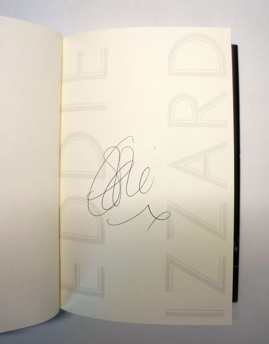 An open book or journal showing a cream-colored inner page with a black handwritten signature in the center. The page has faint large gray lettering in the background, and a dark spine or edge is visible on the right.