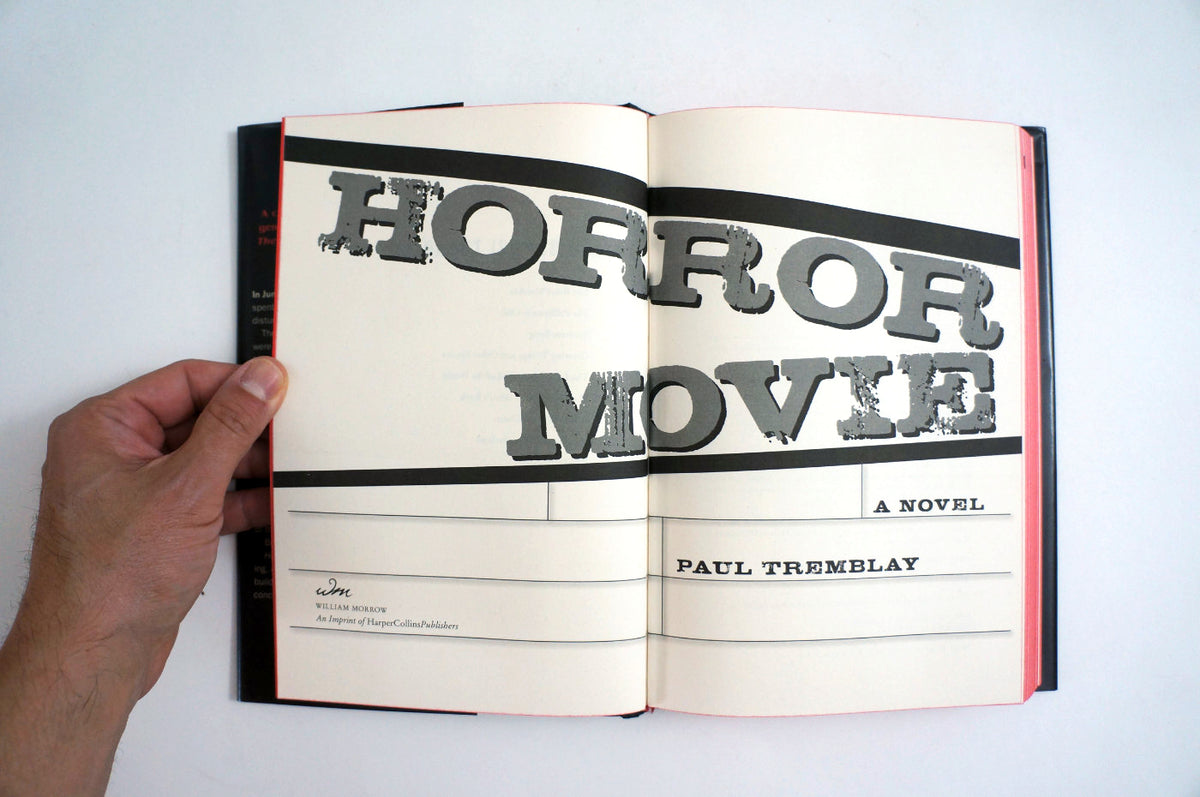 An open book held by a hand, showing the title page of the novel 'Horror Movie' by Paul Tremblay. The pages are off-white with bold black and gray typography, and the book has a dark spine with red accents, set against a plain white background.