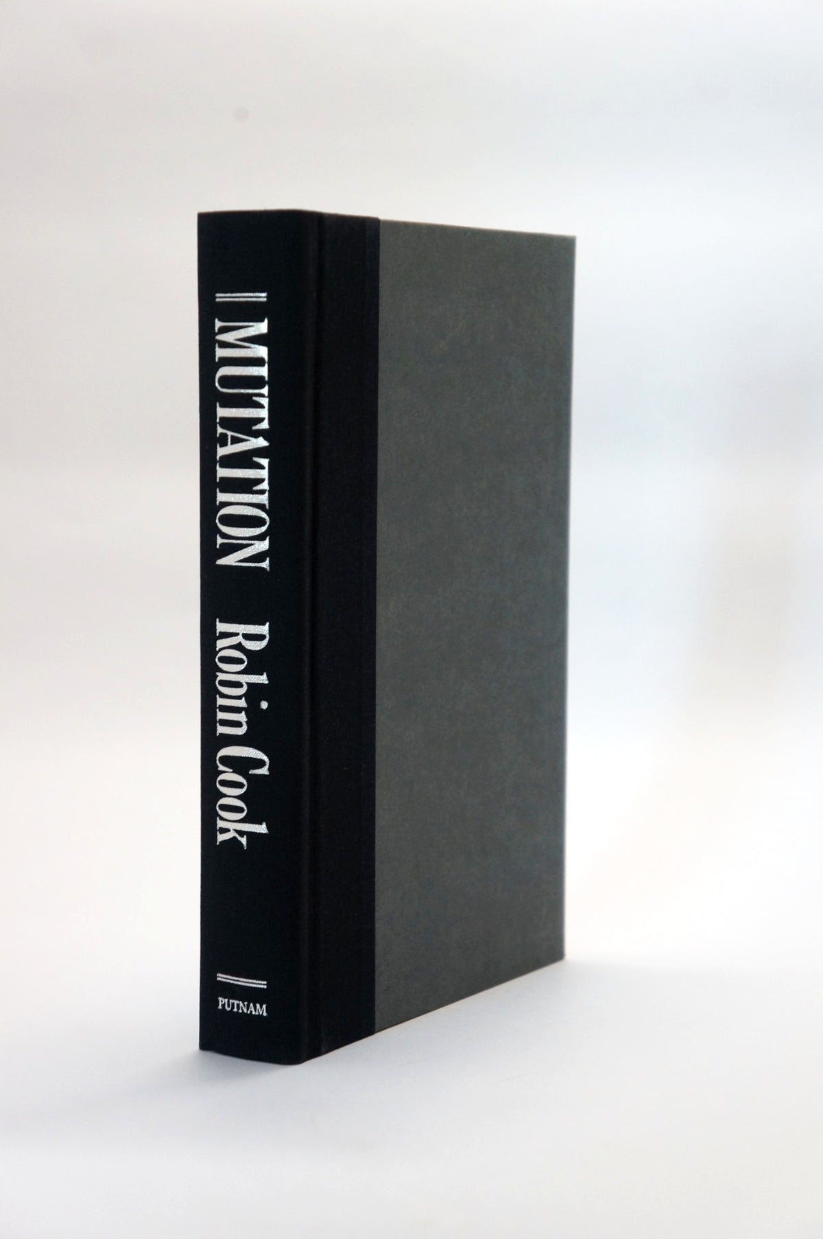 A hardcover book titled 'MUTATION' by Robin Cook, standing upright on a white surface. The book has a black spine with white text and a dark gray textured front cover.