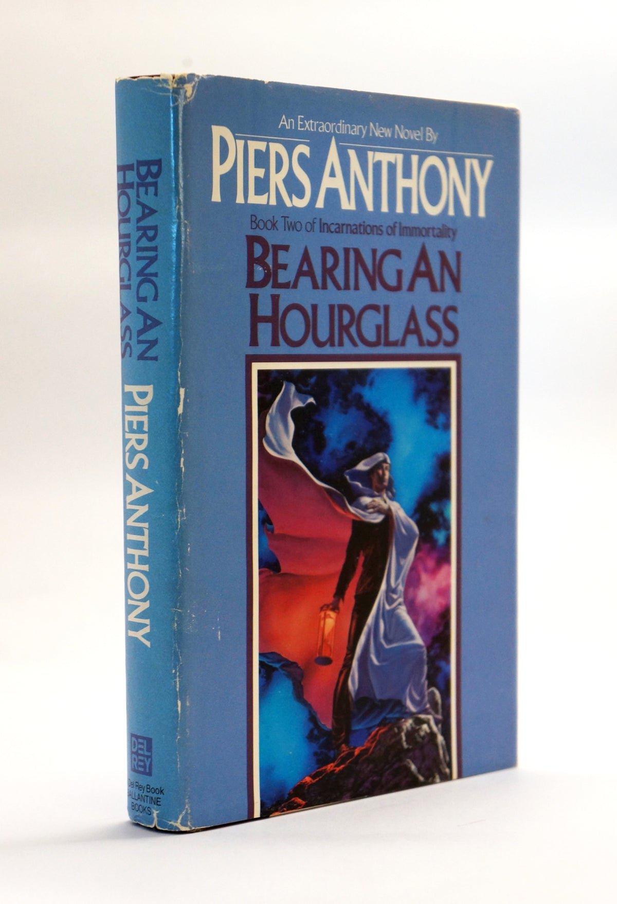 A vintage book of Piers Anthony's 'Bearing an Hourglass' on a white background. The book features a blue cover with a central illustration of a cloaked figure holding a lantern.