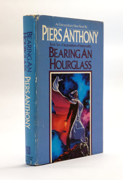 A vintage book of Piers Anthony's 'Bearing an Hourglass' on a white background. The book features a blue cover with a central illustration of a cloaked figure holding a lantern.