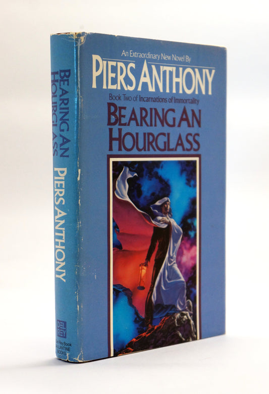 A vintage book of Piers Anthony's 'Bearing an Hourglass' on a white background. The book features a blue cover with a central illustration of a cloaked figure holding a lantern.