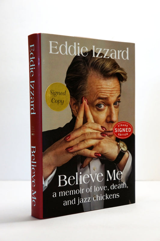 A signed copy of the memoir 'Believe Me' by Eddie Izzard, standing upright on a white surface against a white background. The book features a portrait of the author on the front cover and a dark red spine.