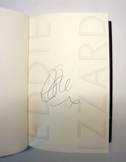 An open book or journal showing a cream-colored inner page with a black handwritten signature in the center. The page has faint large gray lettering in the background, and a dark spine or edge is visible on the right.