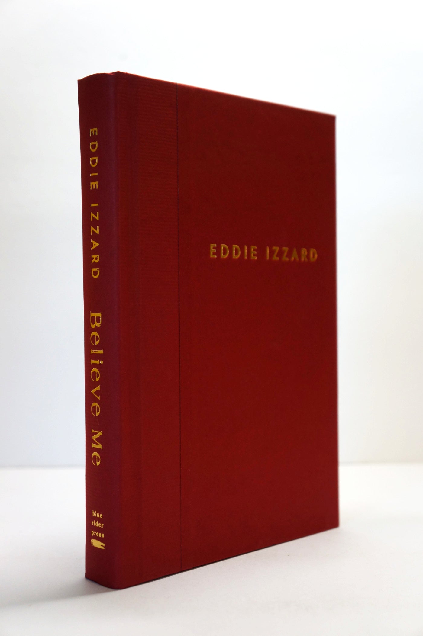 A hardcover book titled 'Eddie Izzard Believe Me' with a textured dark red cover and gold lettering, standing upright on a white surface against a white background.