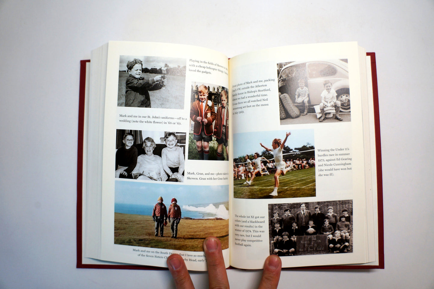 An open hardcover book with printed photographs and text on both pages, held open by two fingers against a plain light background.