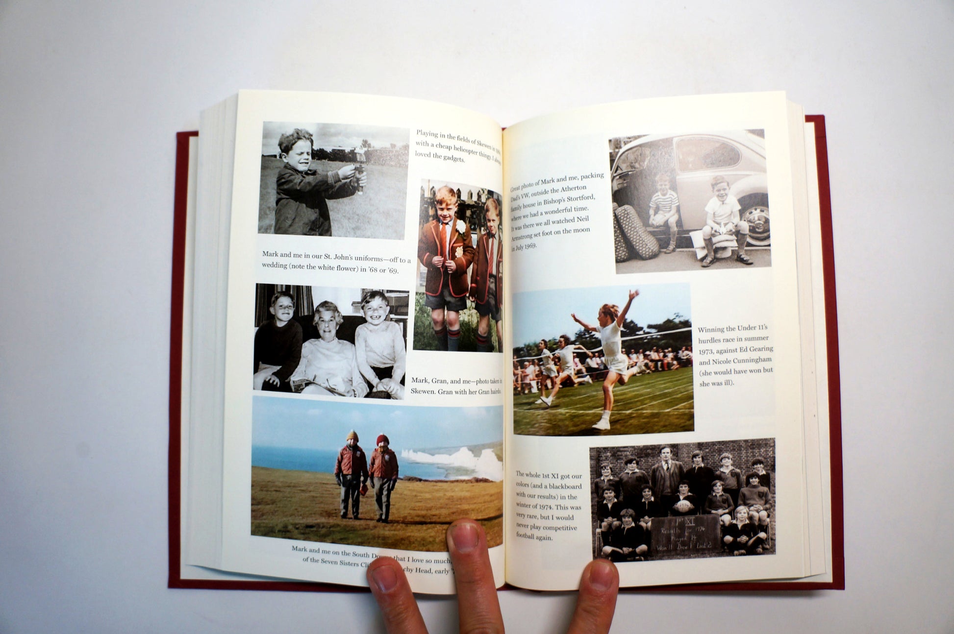 An open hardcover book with printed photographs and text on both pages, held open by two fingers against a plain light background.