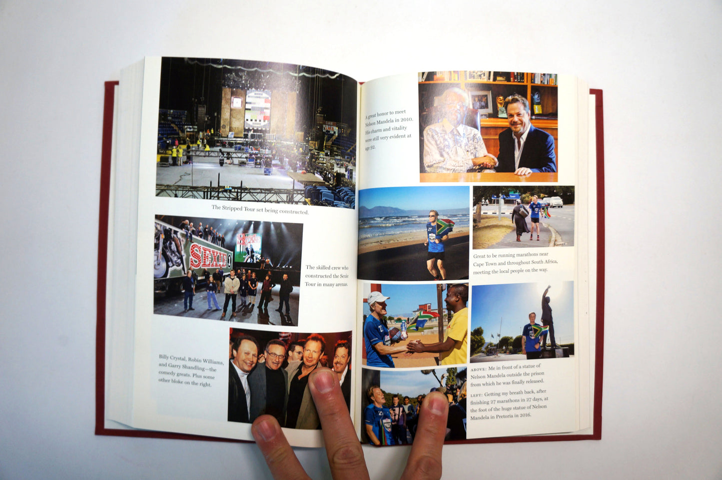 An open hardcover book with red binding and multiple printed photos and text pages, held open by two fingers against a plain white background.
