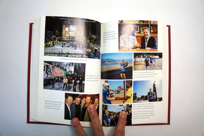 An open hardcover book with red binding and multiple printed photos and text pages, held open by two fingers against a plain white background.