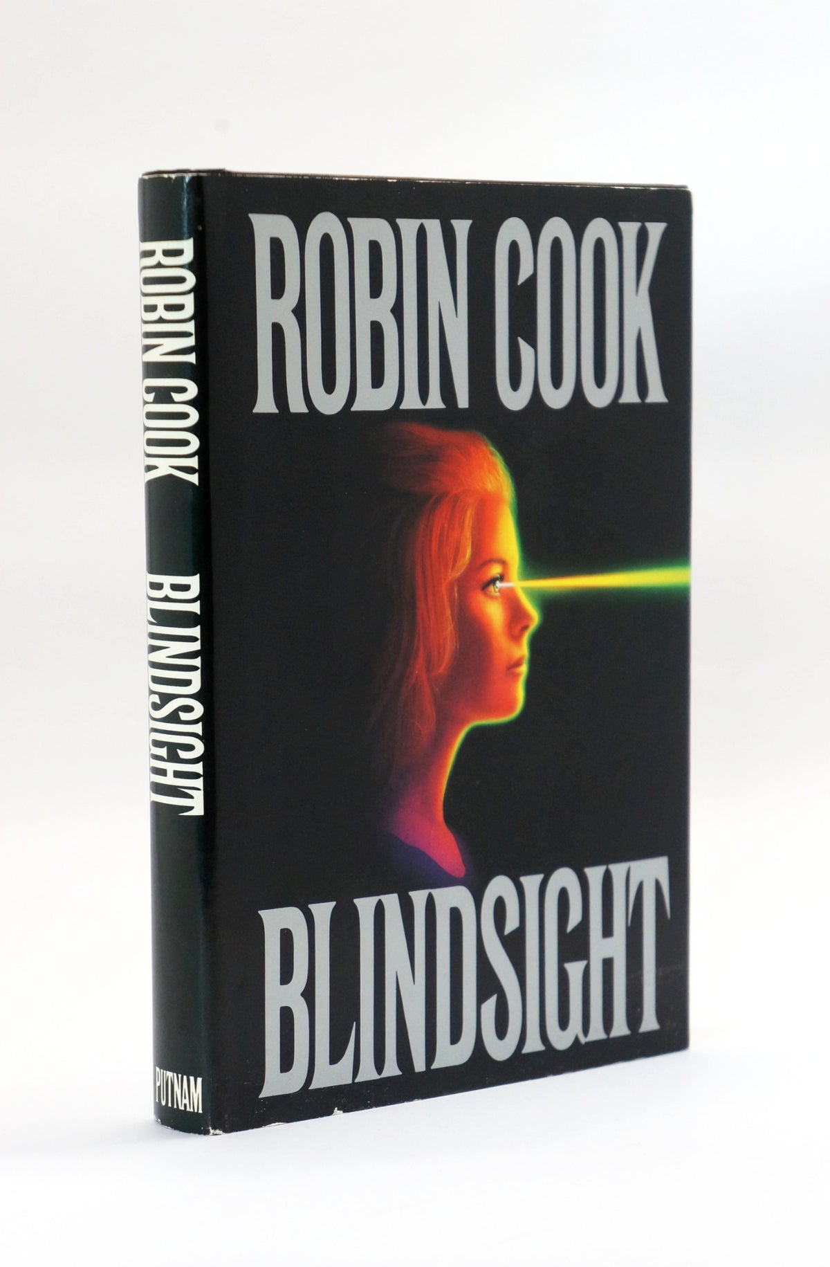 A vintage hardcover book of Robin Cook's 'Blindsight' standing upright on a white background. The cover is black with bold white text and features a stylized profile of a woman's face with a green laser beam projecting from her eye.