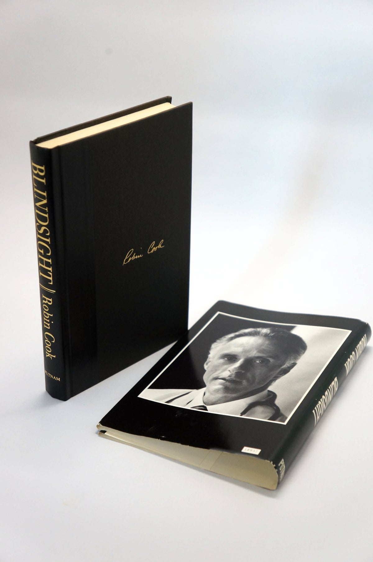 A hardcover book titled 'Blindsight' by Robin Cook, shown standing upright next to its cover laid flat on a light gray background. The cover features a black background with a black-and-white portrait of the author and gold lettering.