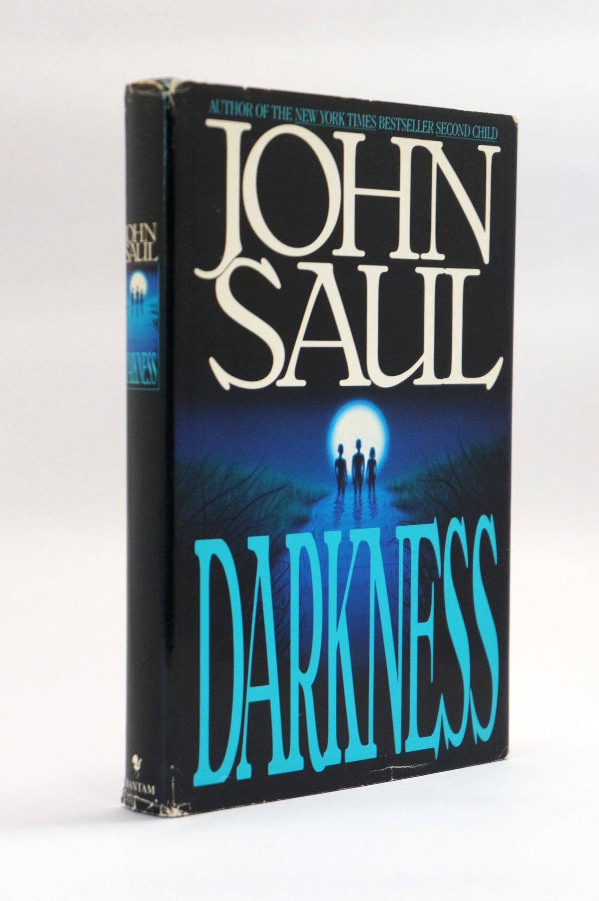 A vintage hardcover book of John Saul's 'Darkness' standing upright on a white background. The cover features a dark blue and black background with a bright moonlit scene and the title in large turquoise letters.