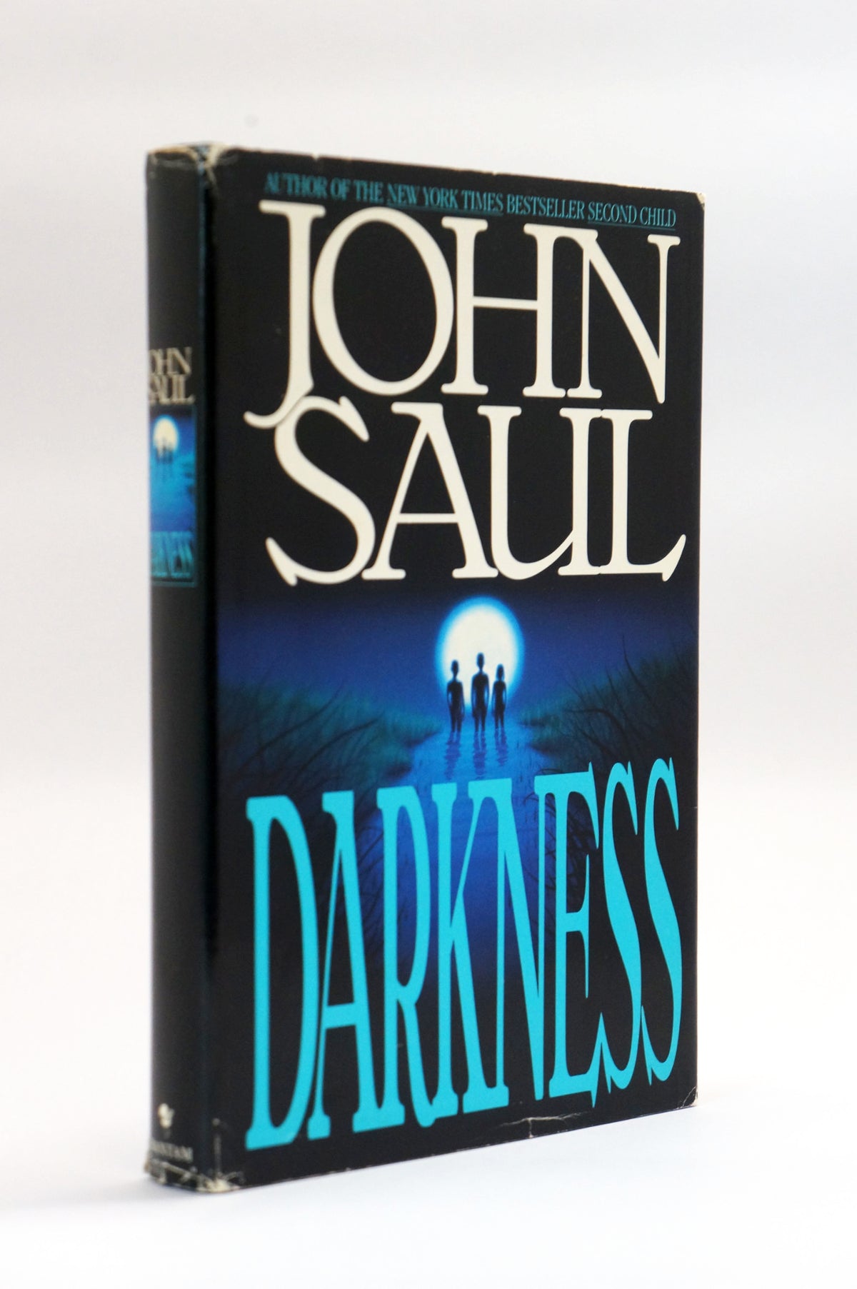 A vintage hardcover book of John Saul's 'Darkness' standing upright on a white background. The cover features a dark blue and black background with a glowing moon and silhouetted figures, and the title 'DARKNESS' is written in large turquoise letters.