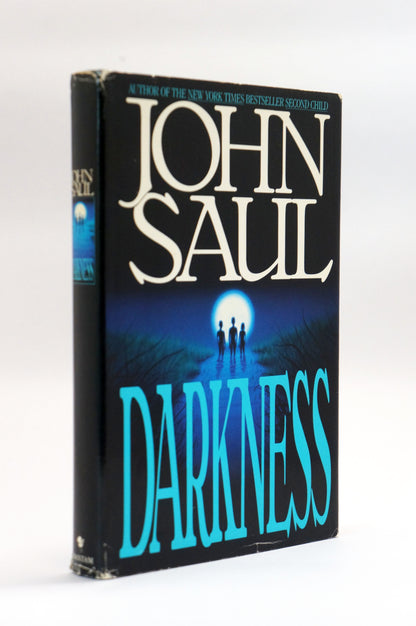 A vintage hardcover book of John Saul's 'Darkness' standing upright on a white background. The cover features a dark blue and black background with a glowing moon and silhouetted figures, and the title 'DARKNESS' is written in large turquoise letters.