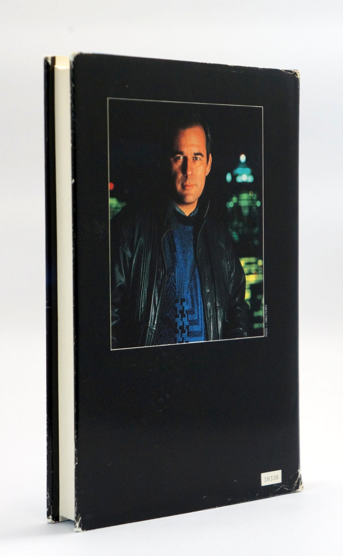 A hardcover book with a black cover featuring a central portrait photo of a man in a dark jacket against a city night background. The book is photographed standing upright on a white surface with a white background.