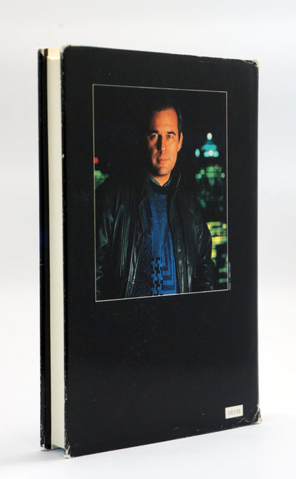 A hardcover book with a black cover featuring a central portrait photo of a man in a dark jacket against a city night background. The book is photographed standing upright on a white surface with a white background.