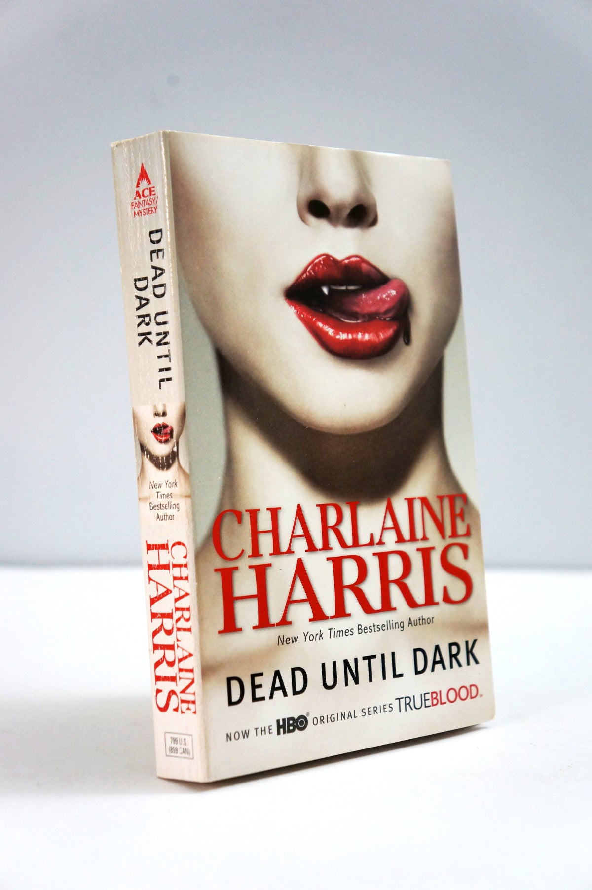 A paperback book of Charlaine Harris's 'Dead Until Dark' standing upright on a light gray background. The cover features a close-up of a woman's face with red lips and a small blood drop, with the title and author name in bold red and black text.