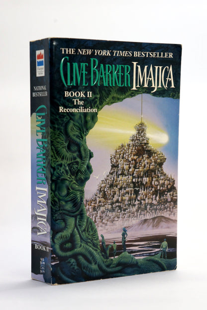 A paperback book of Clive Barker's 'Imajica Book II: The Reconciliation' standing upright on a white background. The cover features a highly detailed fantasy illustration of a massive stone tower under a glowing yellow sky, framed by a large green tree-like creature on the left.