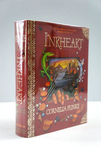 A hardcover book titled 'Inkheart' by Cornelia Funke, shown standing upright at a slight angle. The cover is a deep reddish-brown with ornate gold decorative borders, featuring an illustration of a green dragon and small gold and blue accents.