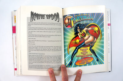 An open book or magazine spread showing a printed article on the left page and a colorful comic-style illustration of a superhero on the right page. A person's fingers are visible holding the bottom edge of the pages against a plain white background.