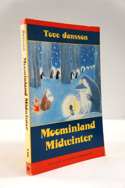 A vintage hardcover book of Tove Jansson's 'Moominland Midwinter' standing upright against a neutral light gray background. The cover features a winter illustration with Moomin characters in a snowy forest, framed by red borders and a dark blue background with yellow title text.