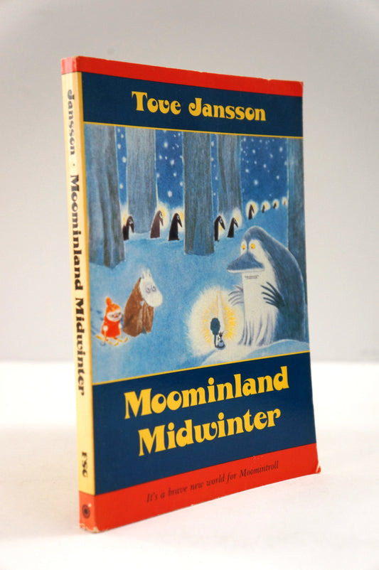 A vintage hardcover book of Tove Jansson's 'Moominland Midwinter' standing upright against a neutral light gray background. The cover features a winter illustration with Moomin characters in a snowy forest, framed by red borders and a dark blue background with yellow title text.