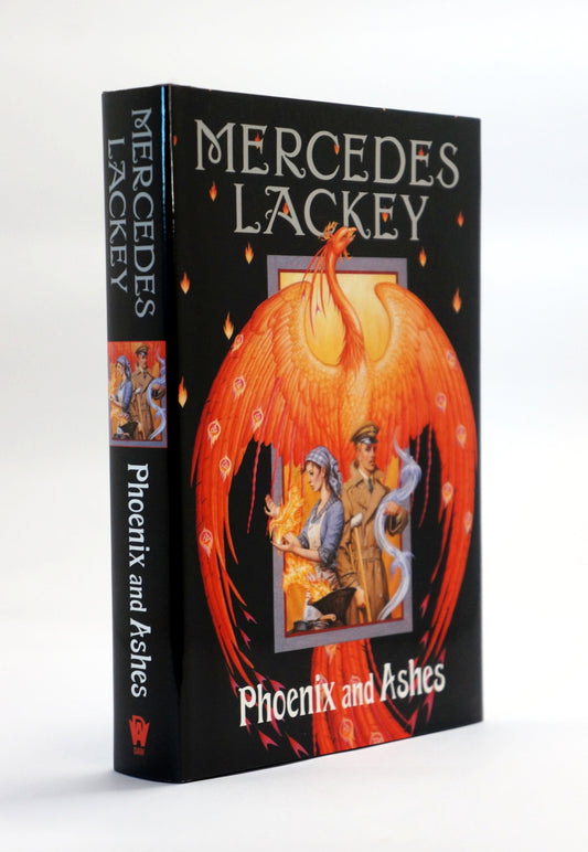A hardcover book titled 'Phoenix and Ashes' by Mercedes Lackey, standing upright at a slight angle. The book cover is predominantly black with a large fiery phoenix illustration in orange and red tones, and the title and author name are printed in white text.
