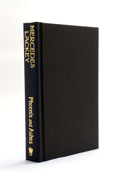 A black hardcover book standing upright at a slight angle, showing the spine with gold text reading 'MERCEDES LACKEY' and 'Phoenix and Ashes,' on a clean white background.