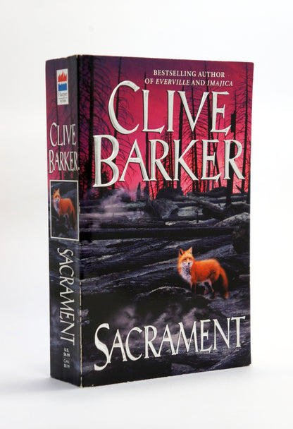 A paperback of Clive Barker's novel 'Sacrament' standing upright on a white background. The cover features a dark, eerie forest scene with a small orange fox in the foreground and the author's name in large white letters.