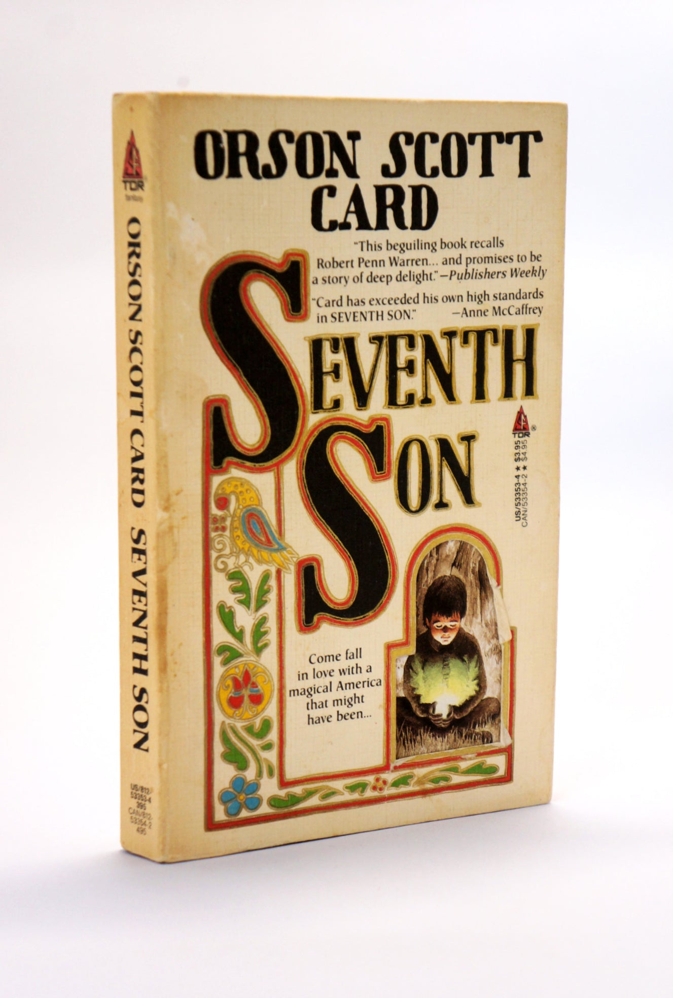 A vintage paperback book of Orson Scott Card's 'Seventh Son' standing upright on a white background. The cover is a warm beige with large stylized black and brown lettering for the title, and a small illustration of a boy in a green cloak in a window frame.