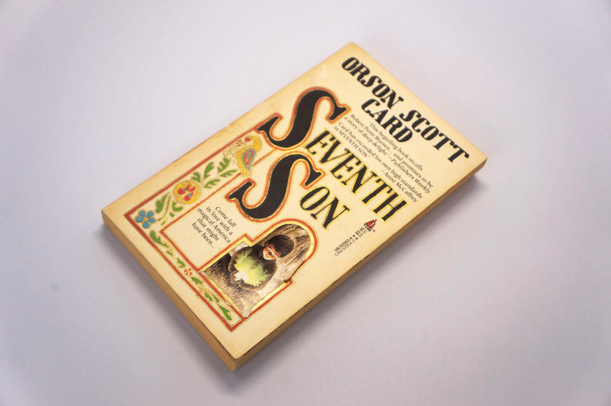 A vintage paperback book of Orson Scott Card's 'Seventh Son' resting on a plain white surface, angled diagonally. The book cover is a warm cream/beige color with large black stylized lettering for the title and a small vintage-style portrait illustration in the lower-left corner.