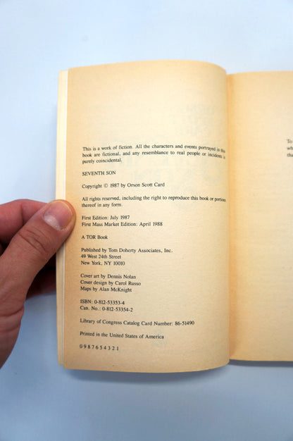 A hand holding an open vintage paperback book, showing the printed copyright and publisher information on the left page against a light blue background.