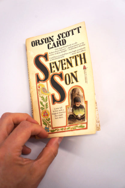 A hand holding a vintage paperback book of Orson Scott Card's 'Seventh Son' against a plain white background.