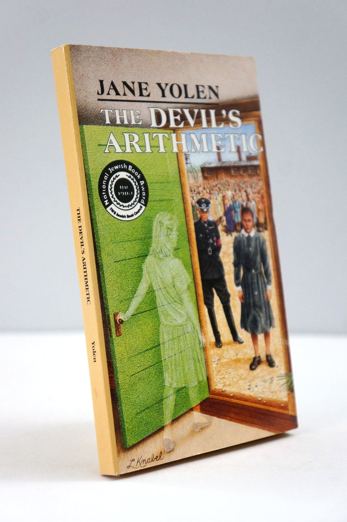 A paperback titled "The Devil's Arithmetic" by Jane Yolen, standing upright at a slight angle against a plain light gray background. The cover features a tan border with a central illustration split into a green door opening and a schoolhouse scene with children and adults.
