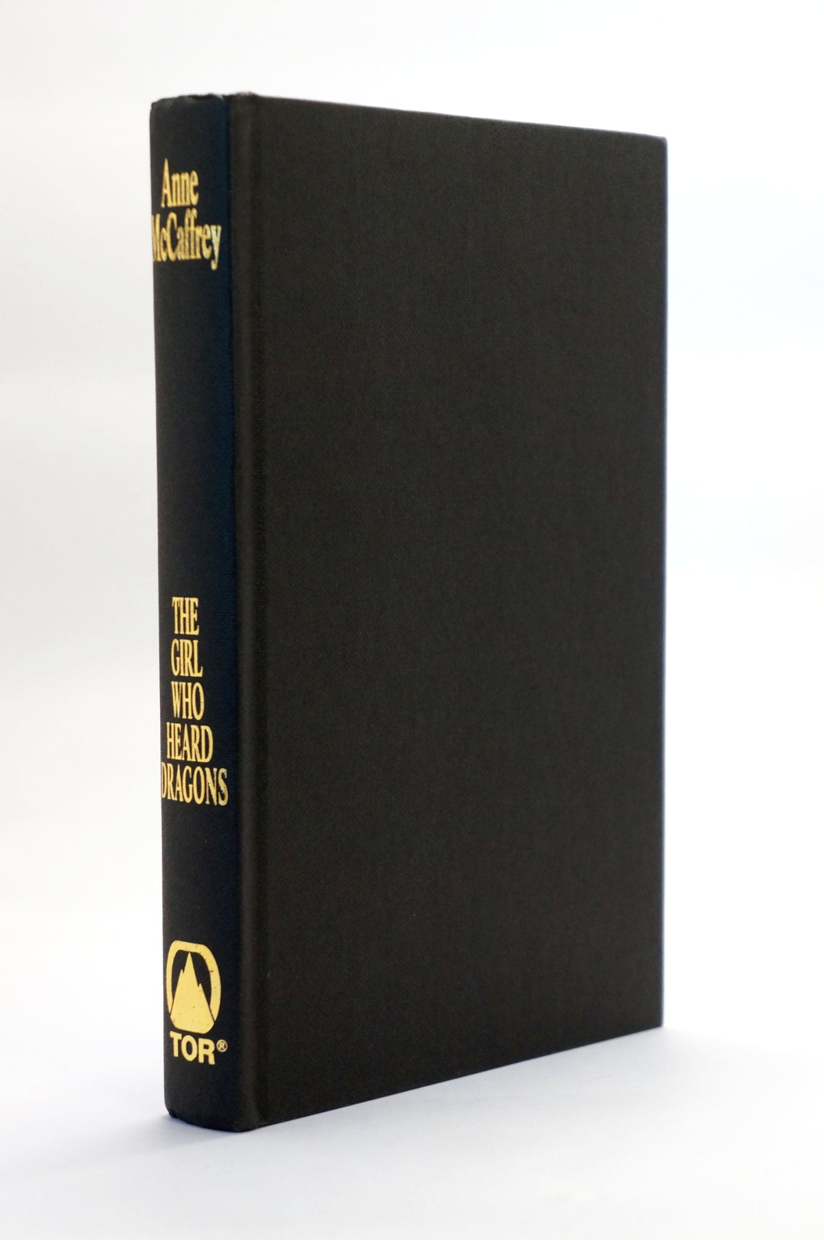 A hardcover book standing upright at a slight angle, showing its black cloth-protected front cover and dark blue spine with gold lettering. The spine reads 'Anne McGillrey', 'THE GIRL WHO Heard DRAGONS', and the TOR logo.