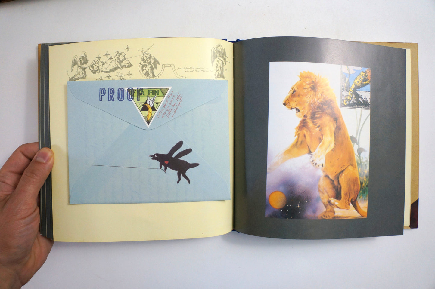 An open book or album spread held by a hand, showing printed pages with a light blue envelope labeled 'PROOF EN' and a black rabbit silhouette, alongside a colorful lion illustration on the right page.