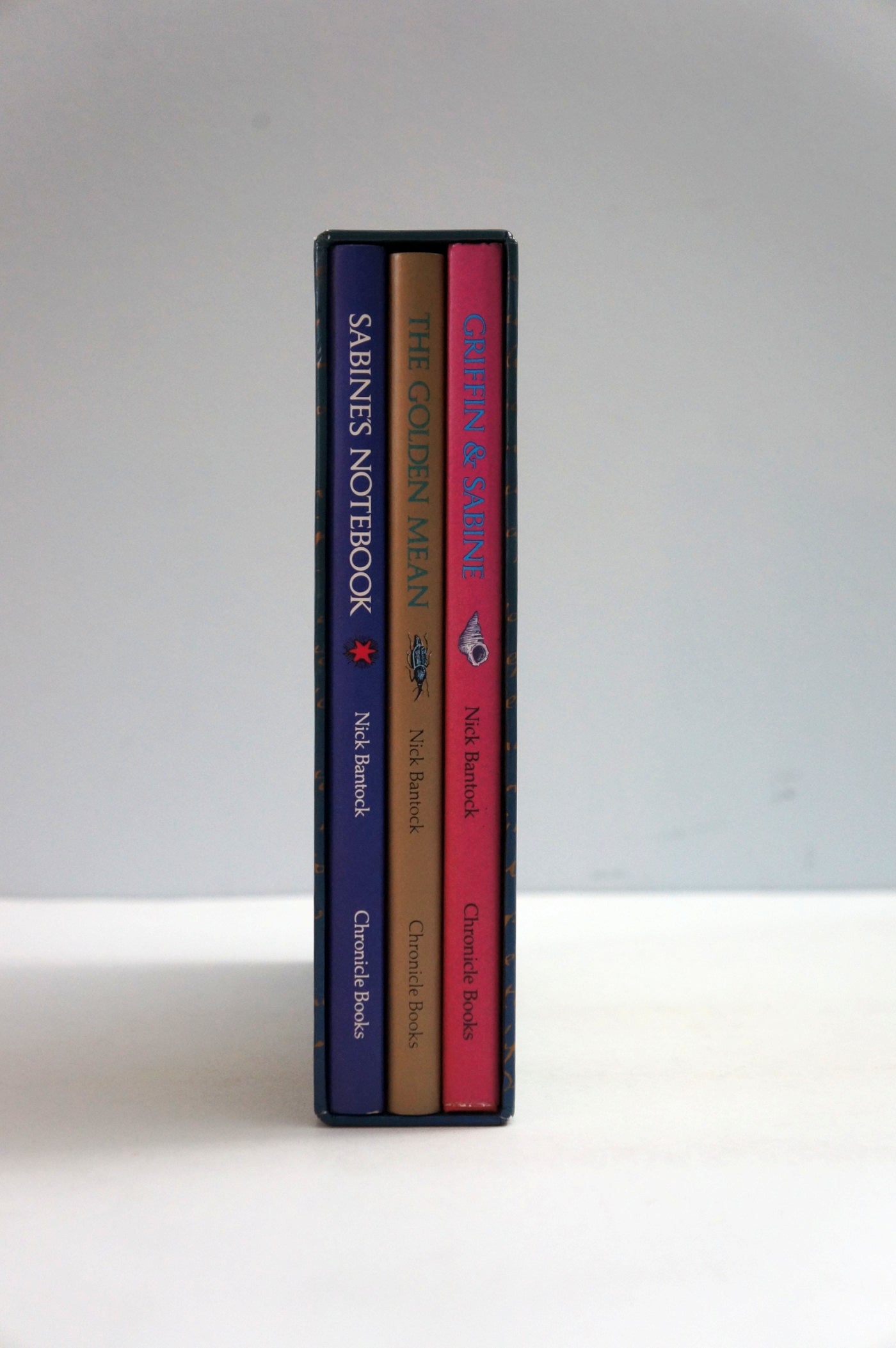 Three slim notebooks standing upright side by side on a white surface against a light gray background. The spines show the titles 'SHRINES NOTEBOOK', 'THE GOLDEN MEAN', and 'Crimson & Clover', with dark blue, tan, and pink covers.