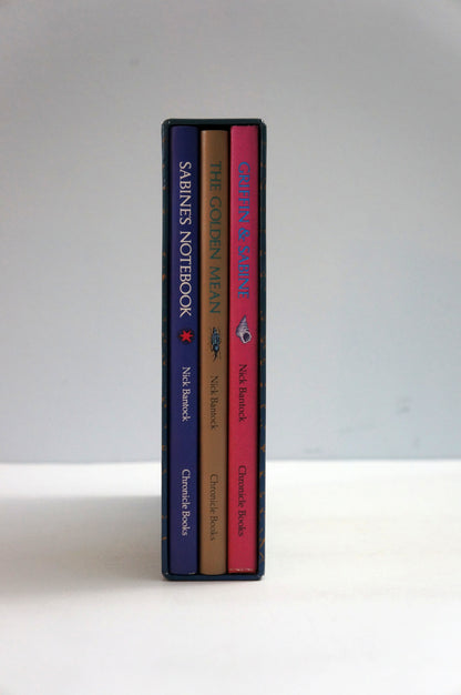 Three slim notebooks standing upright side by side on a white surface against a light gray background. The spines show the titles 'SHRINES NOTEBOOK', 'THE GOLDEN MEAN', and 'Crimson & Clover', with dark blue, tan, and pink covers.