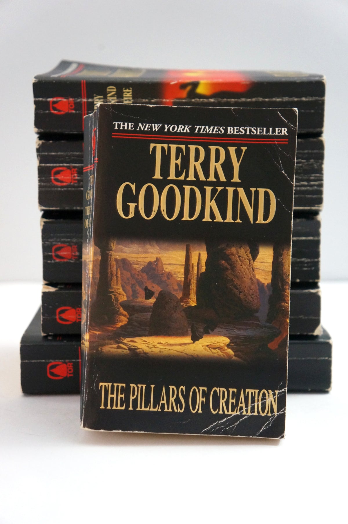 A stack of hardcover books with the front book titled "The Pillars of Creation" by Terry Goodkind prominently displayed. The book cover is black with large gold lettering and a desert landscape illustration.
