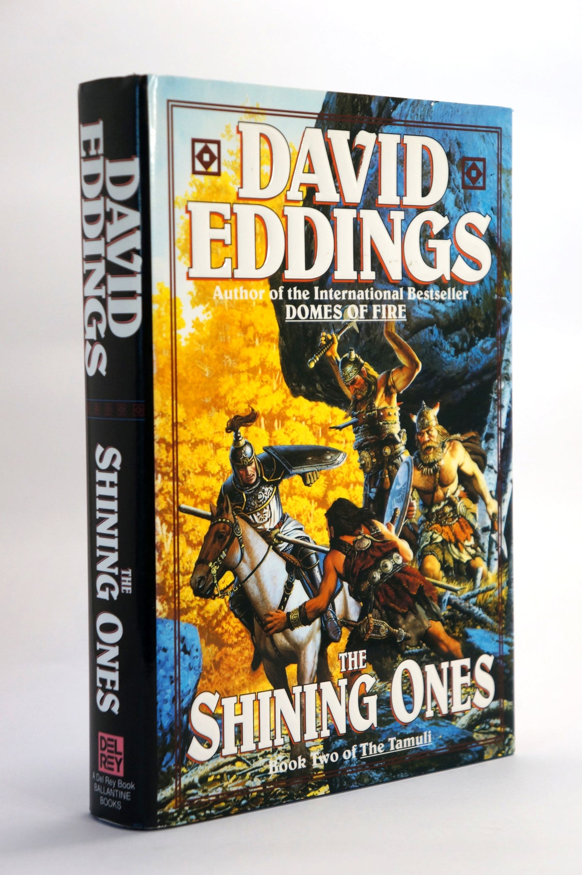 A hardcover book of David Eddings' 'The Shining Ones' standing upright at a slight angle. The cover features a dramatic fantasy illustration with a warrior on a white horse against a fiery yellow and blue background, with the author's name in large white letters at the top.