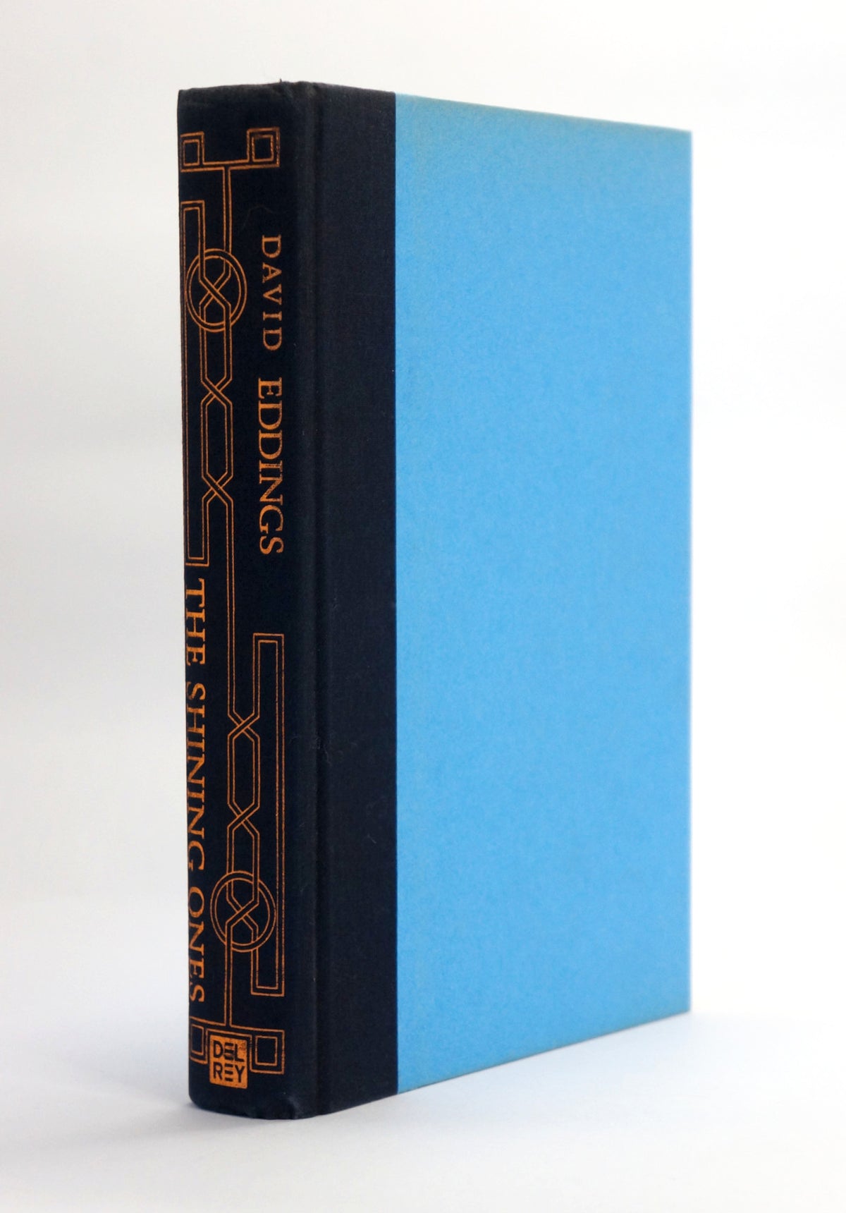 A hardcover book standing upright at a slight angle, showing a dark navy blue spine with gold decorative line art and the title 'The Shining Ones' by David Eddings, next to a light blue front cover, on a white background.