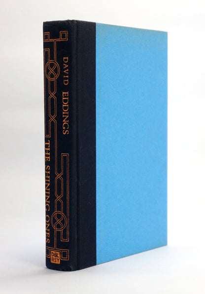 A hardcover book standing upright at a slight angle, showing a dark navy blue spine with gold decorative line art and the title 'The Shining Ones' by David Eddings, next to a light blue front cover, on a white background.