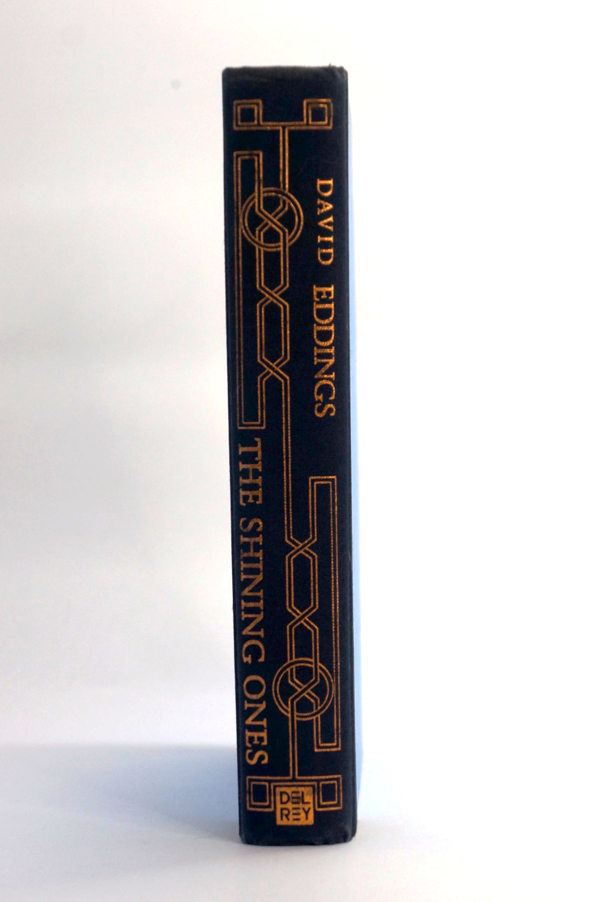 The spine of a hardcover book titled 'The Shining Ones' by David Eddings, featuring a dark navy blue cover with gold geometric line art and gold lettering, photographed against a plain white background.